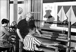 An elderly white woman stands over a young white boy who smiles while he operates a large loom. 